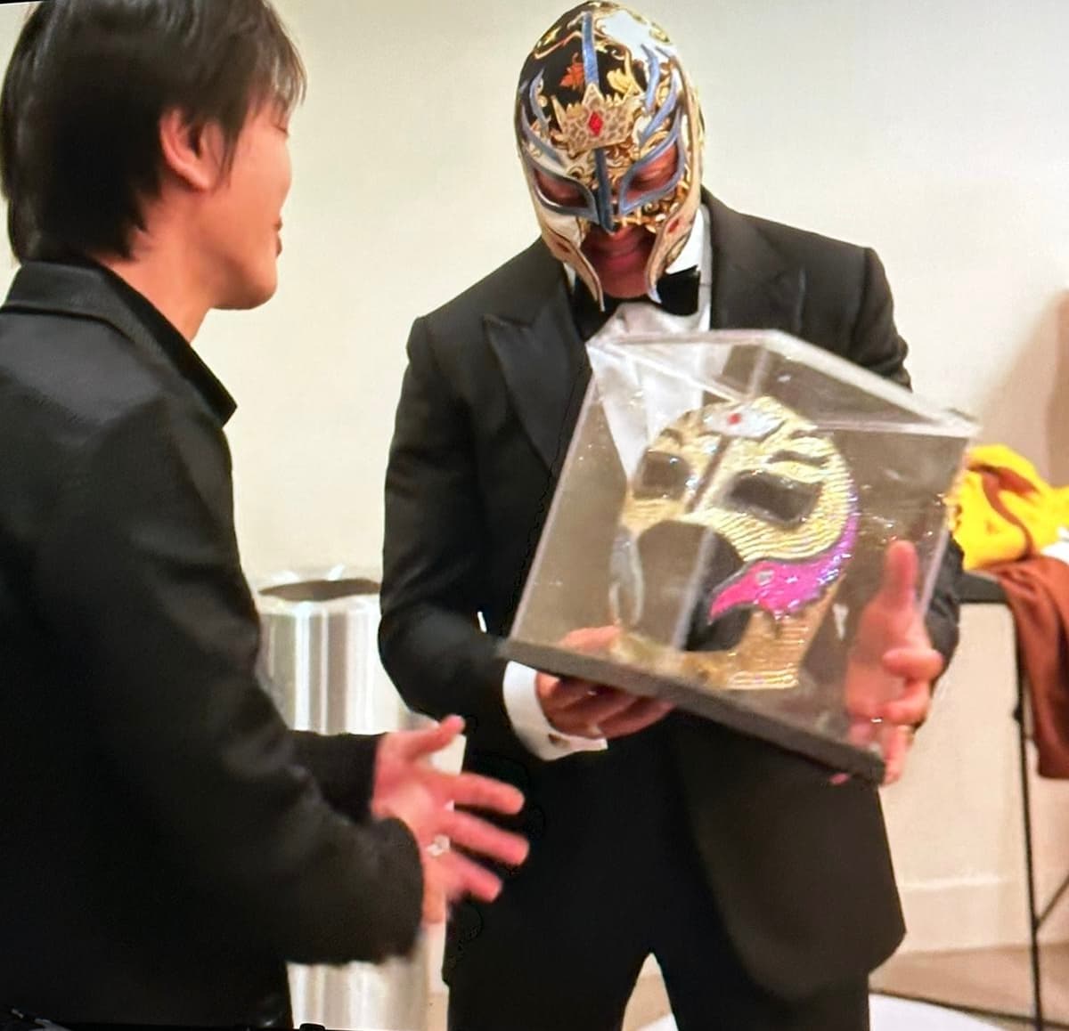 Rey Mysterio in a tuxedo presents a luchador mask in a clear display case.