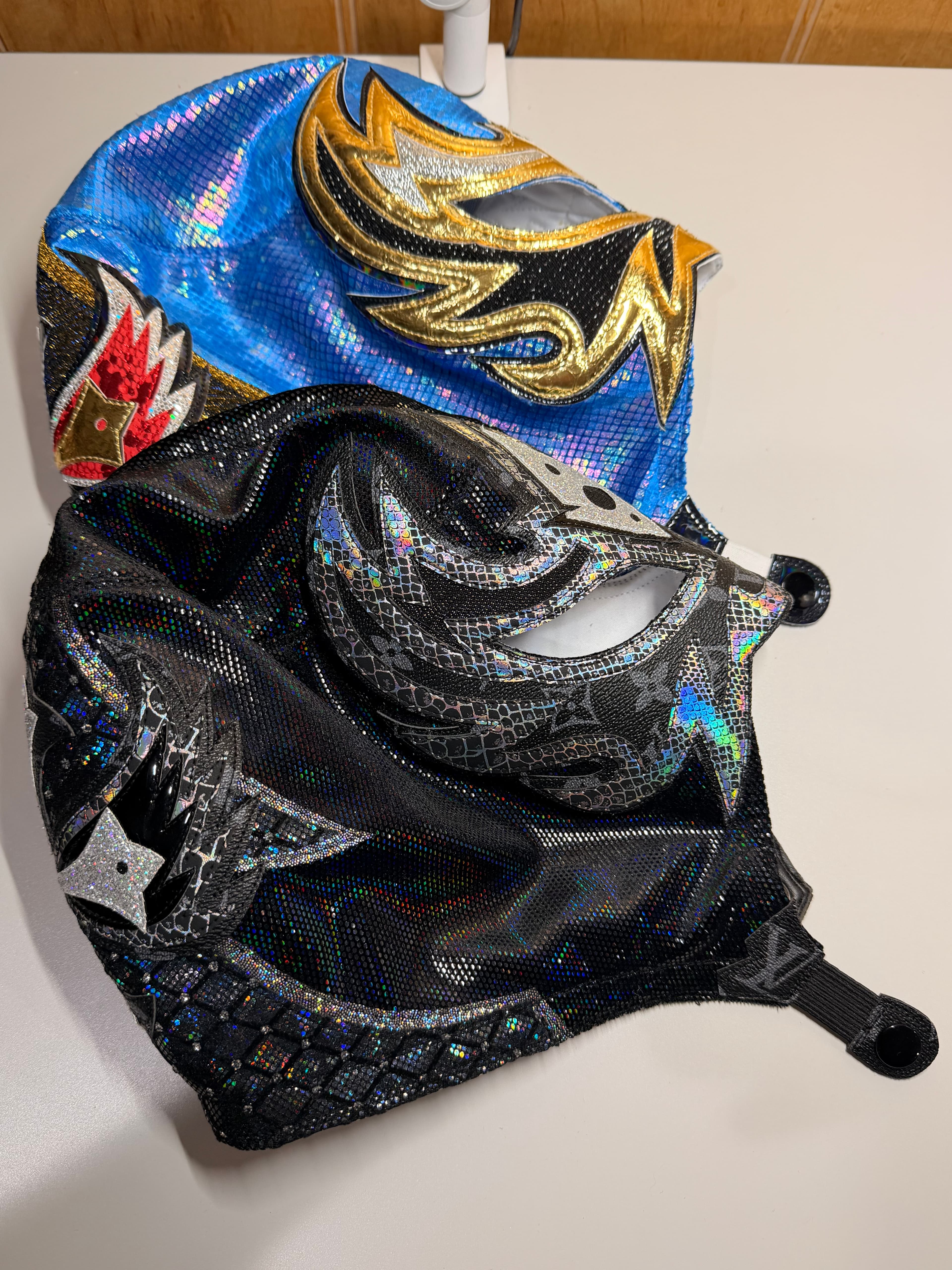 Two iridescent Lucha Libre masks, one blue with gold flames, one black with silver patterns.