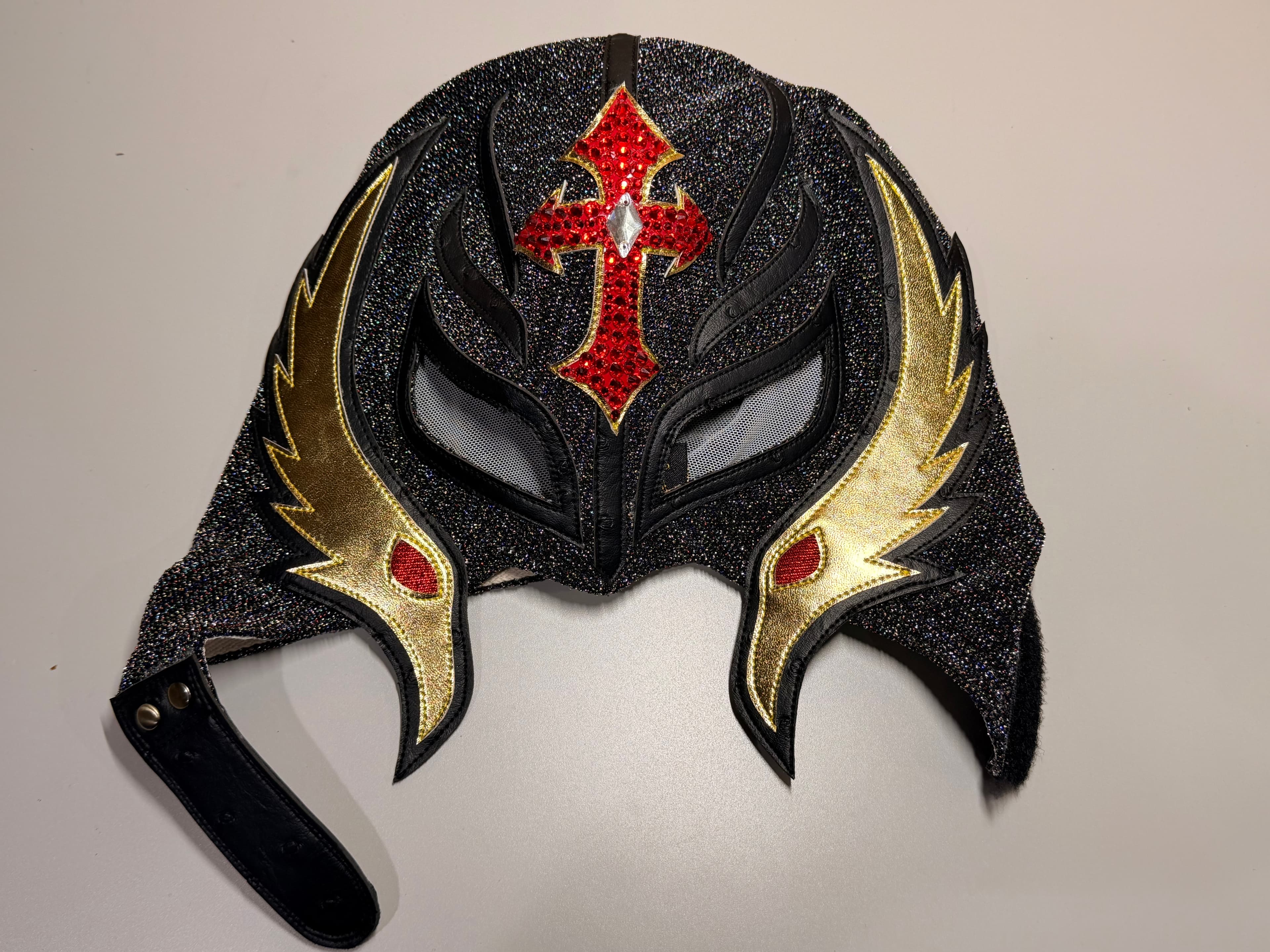Black glittery lucha libre mask with a red jeweled cross and gold wing-like patterns.