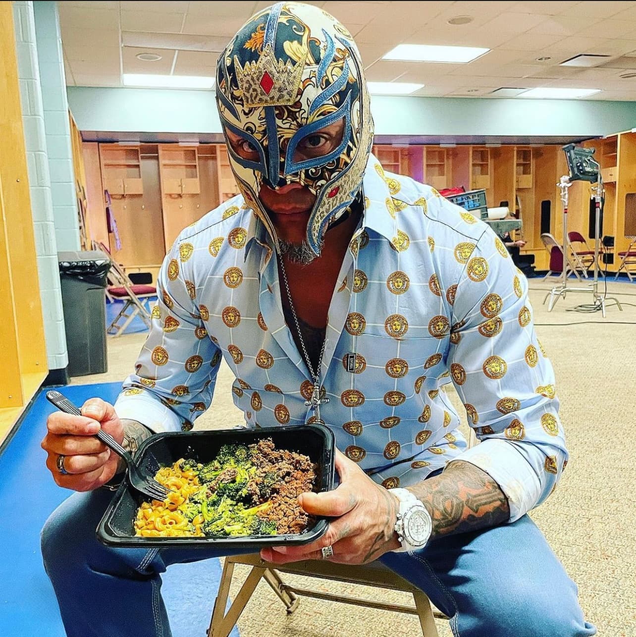 Rey Mysterio in luchador mask and patterned shirt, eating a meal in a locker room.