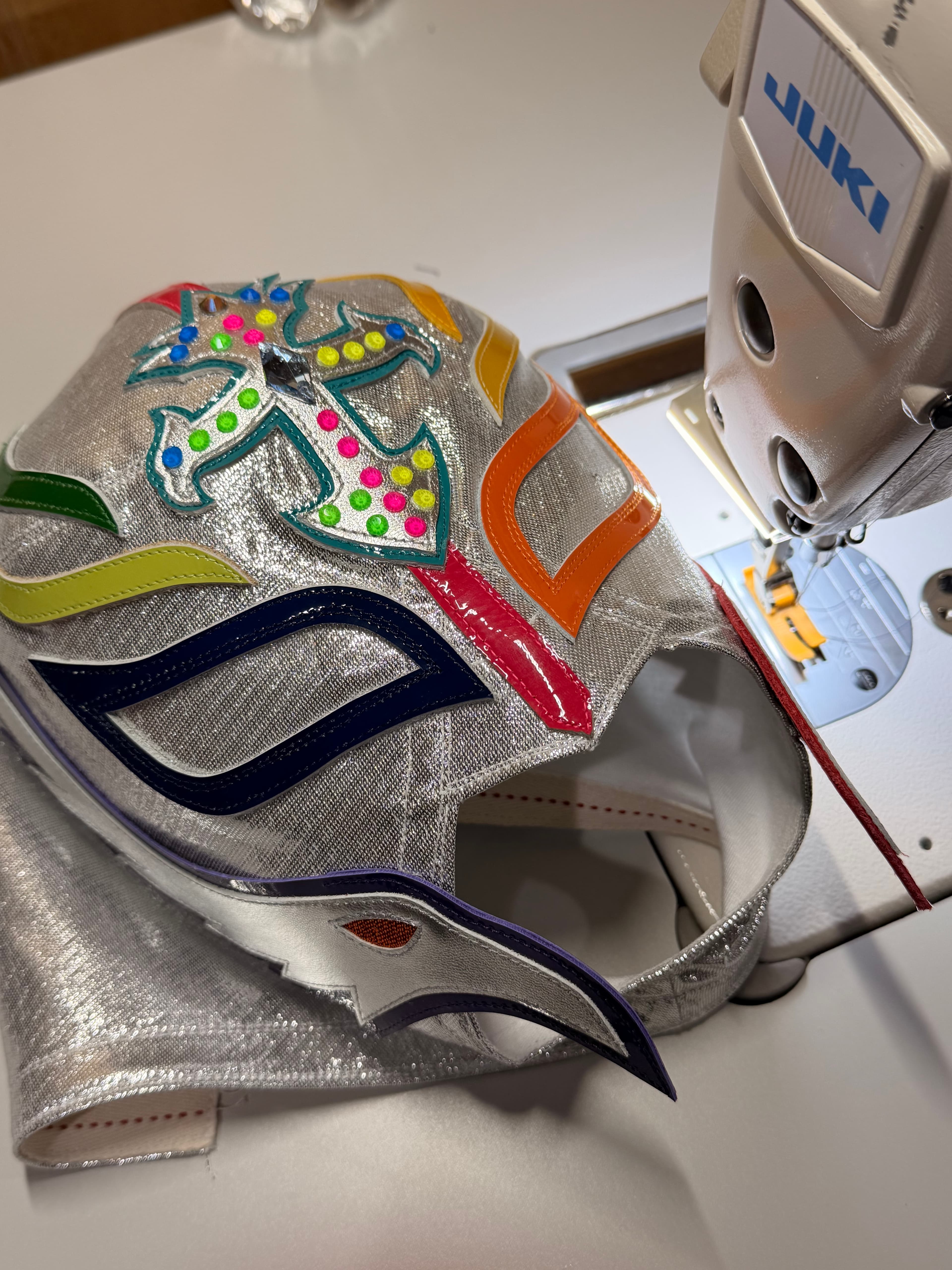 Vibrant silver luchador mask with colorful appliqués under a white Juki industrial sewing machine.