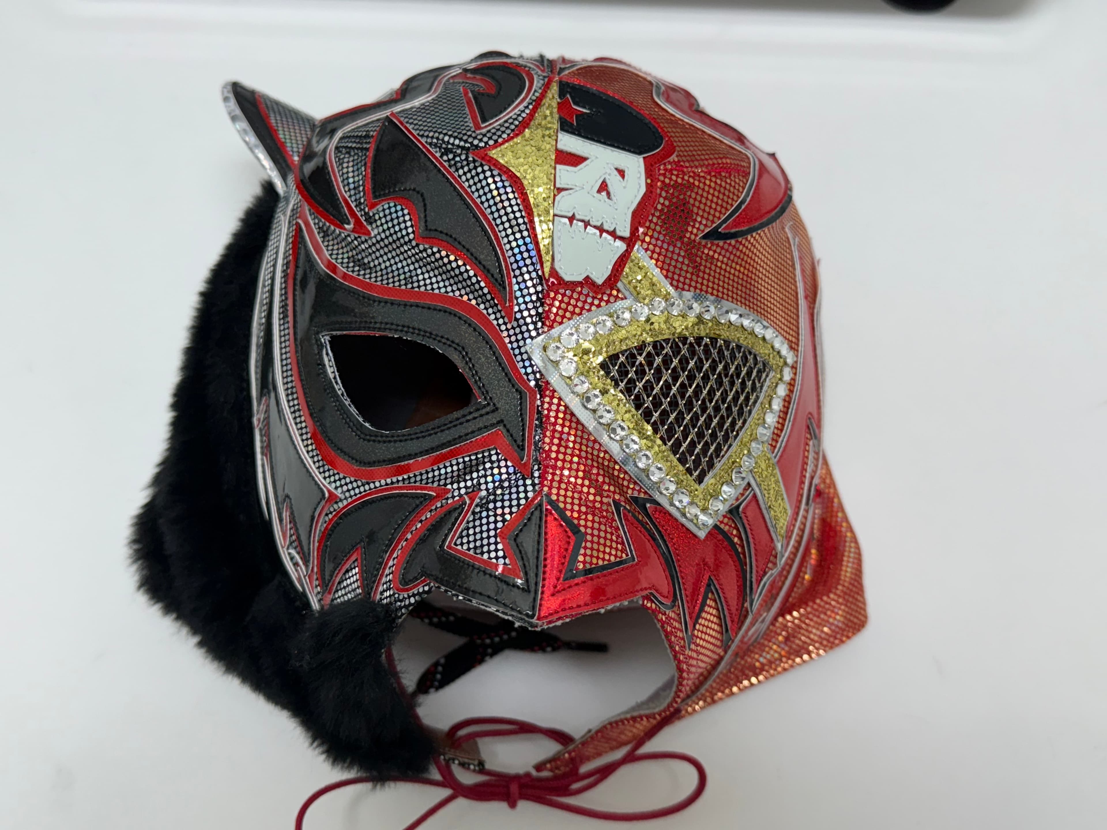 Ornate red and black Lucha Libre mask with silver patterns, skull emblem, and black fur.
