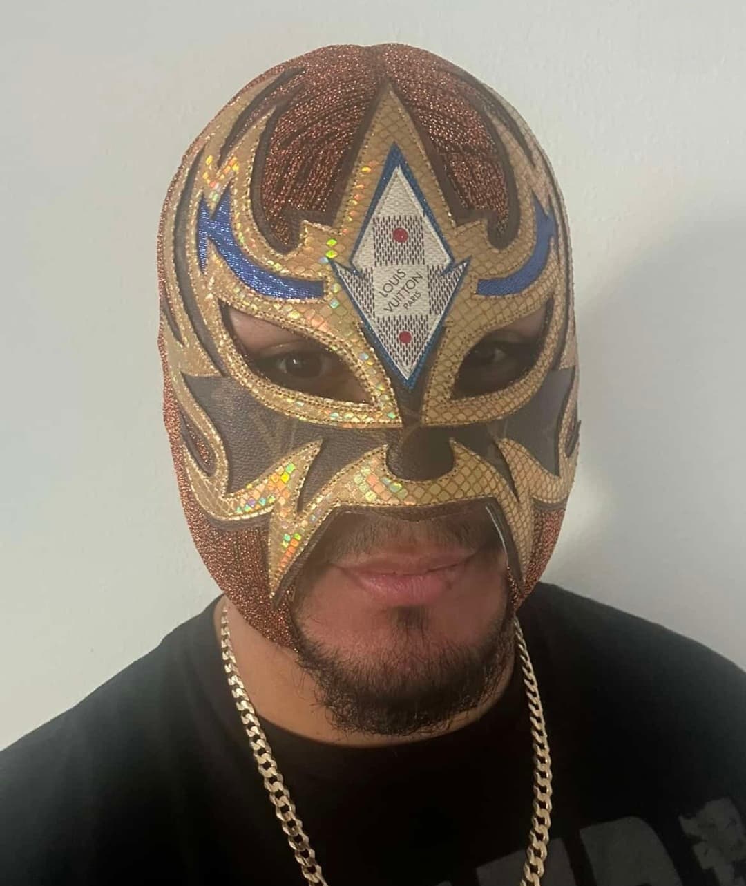 Man wearing a brown and gold luchador mask featuring a Louis Vuitton logo on forehead.
