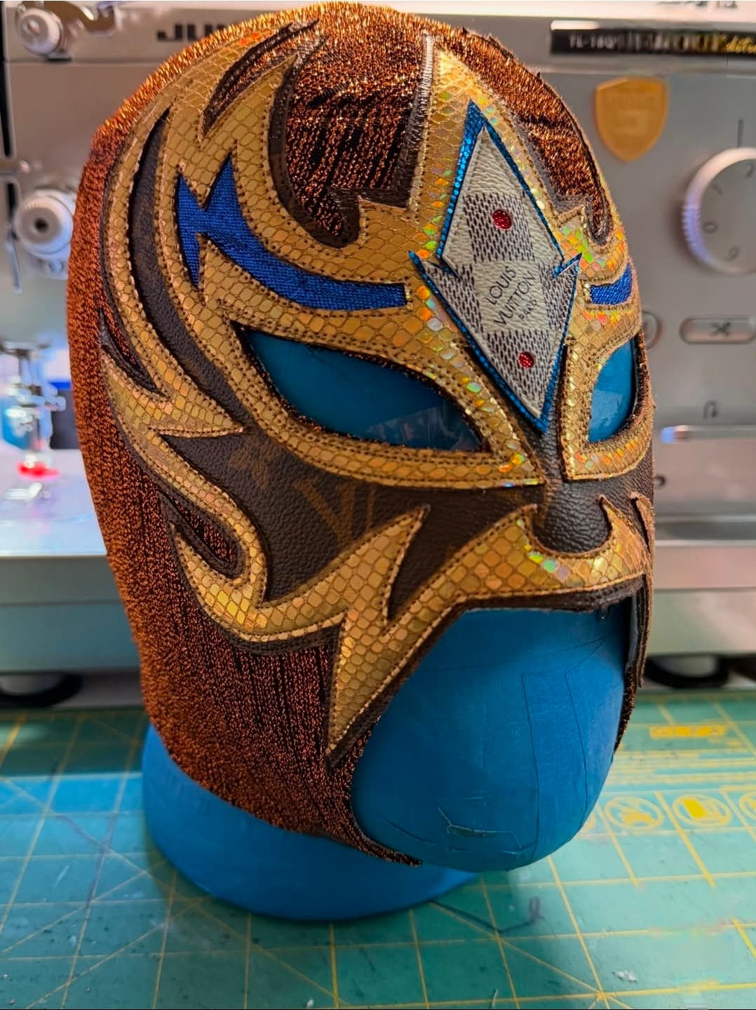 Bronze and gold Lucha Libre mask with Louis Vuitton monogram on a blue mannequin.