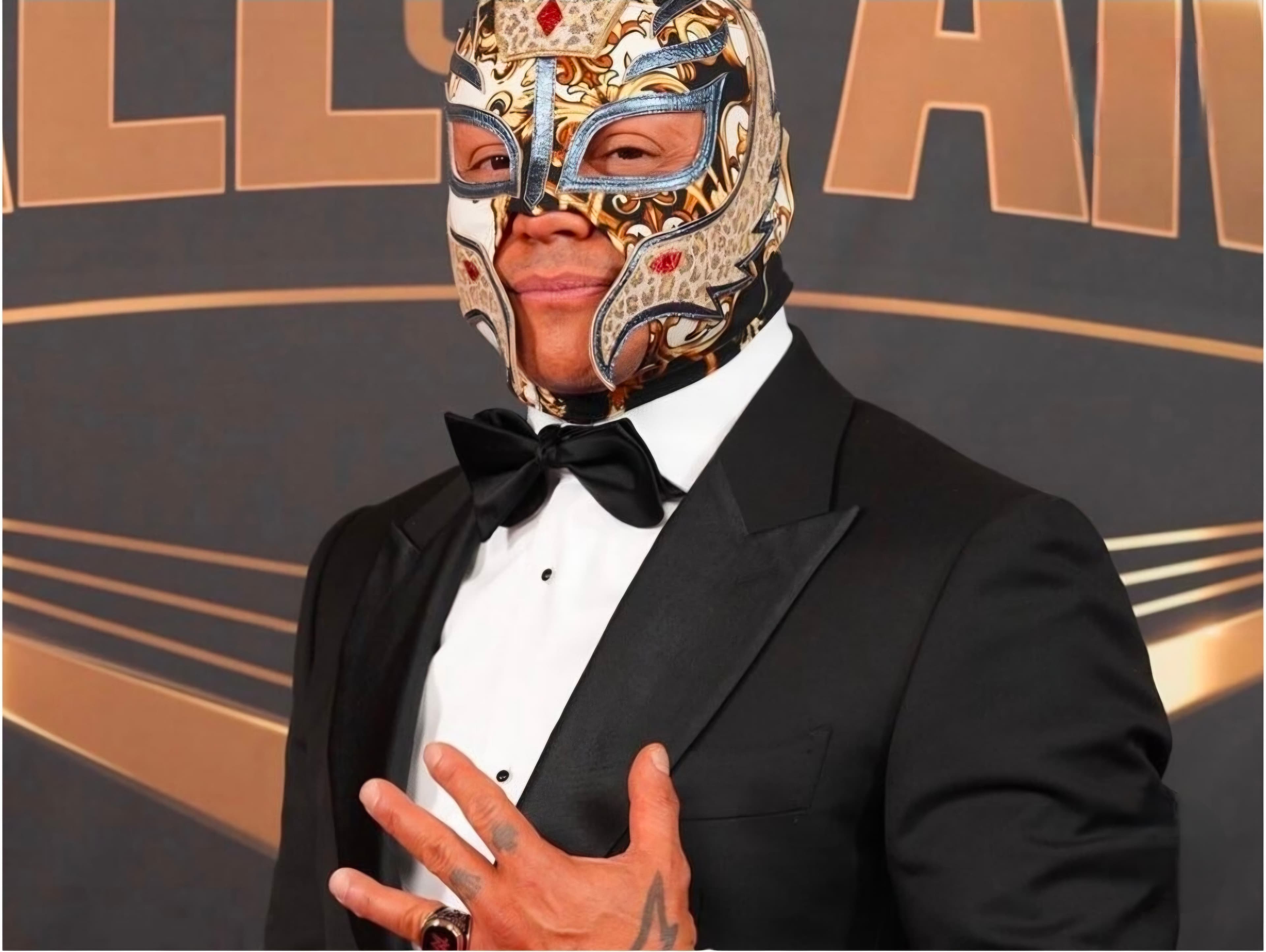 Rey Mysterio wearing an ornate gold and white wrestling mask with a black tuxedo.
