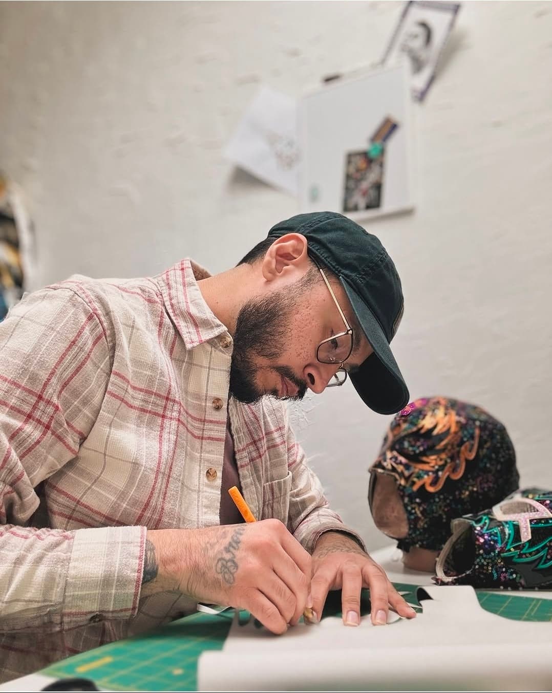 Bearded man in a plaid shirt and cap carefully crafts a colorful lucha libre mask.