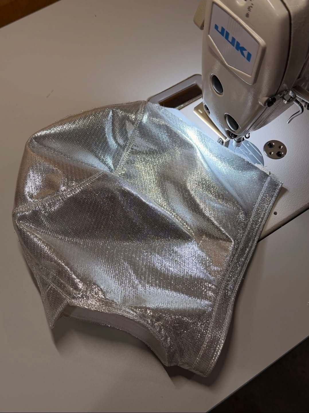 Shiny silver metallic fabric being sewn on a white Juki sewing machine under bright light.