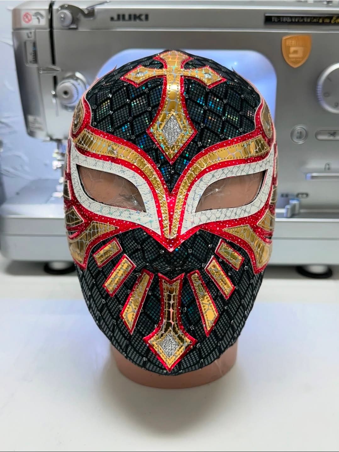 Intricate black Lucha Libre mask with gold, red, and white patterns on a mannequin head.