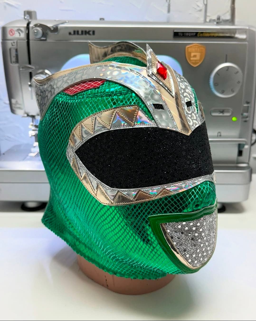 Green scaly luchador mask with silver accents and black visor, displayed before a sewing machine.
